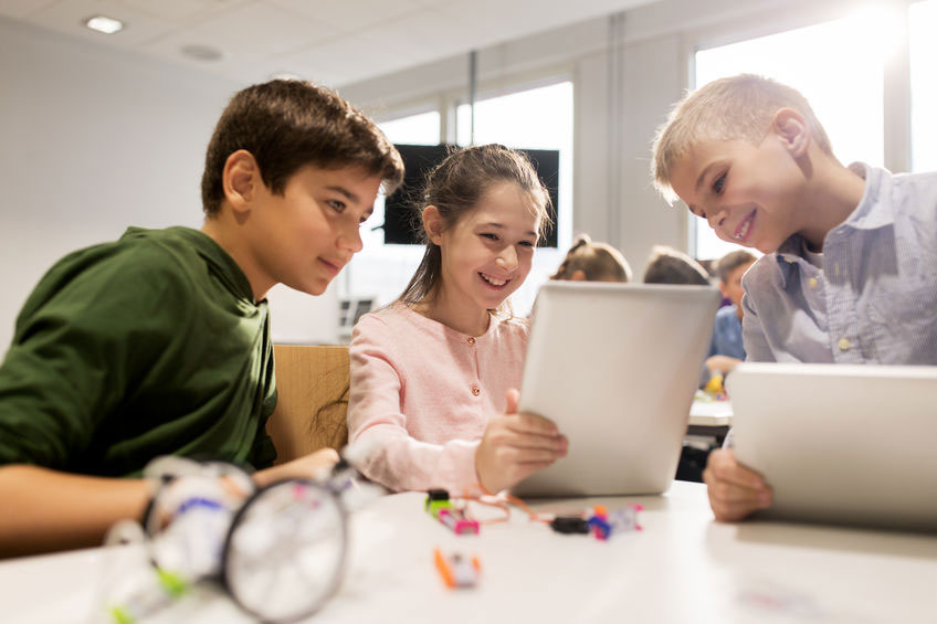 drie schoolkinderen werken op laptops. Op de achtergrond een groot beeldscherm.