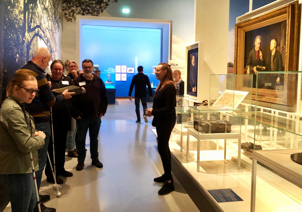 Blinde en slechtziende museumbezoekers luisteren naar de rondleider en betasten een object, vergelijkbaar als in het schilderij waar ze voor staan.