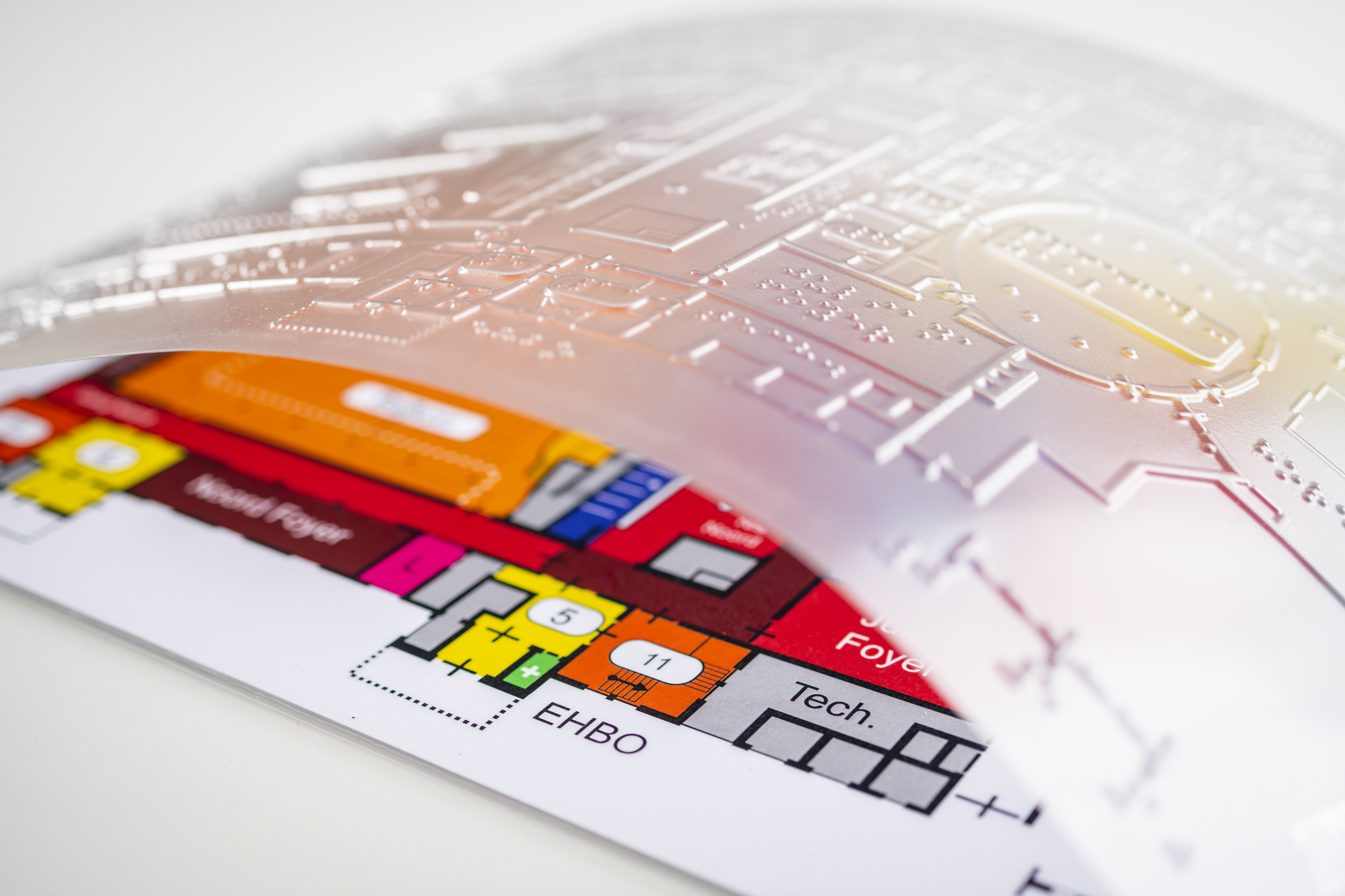 Een plattegrond voorzien van een kaart in felle kleuren en grootletter en daar bovenop een doorzichtig folie met de plattegrond in reliëf en braille
