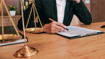 persoon aan bureau met pen in de hand en tekst op papier en een weegschaal